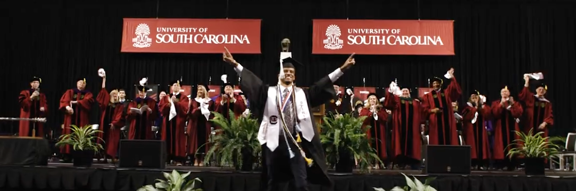 Male student celebrating his graduation day on the graduation stage surrounded by faculty members who are cheering him on.