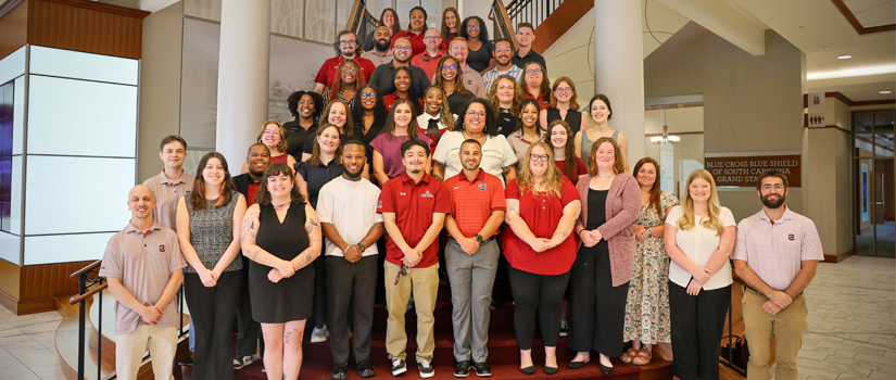 Group photo of the Residence Life staff standing on a staircase inside a bright, modern campus building. Approximately 40 staff members are arranged in several rows, smiling and facing the camera. The group includes individuals wearing professional attire in university colors, with a spacious lobby, large columns, and upper-level balconies visible in the background, creating a welcoming and professional atmosphere.