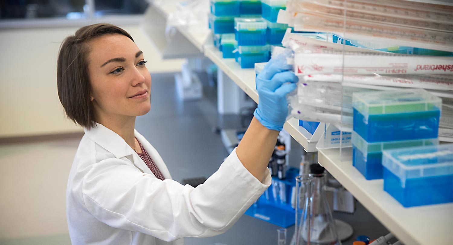 Researcher wearing a lab coat reaching for equipment on a shelf.
