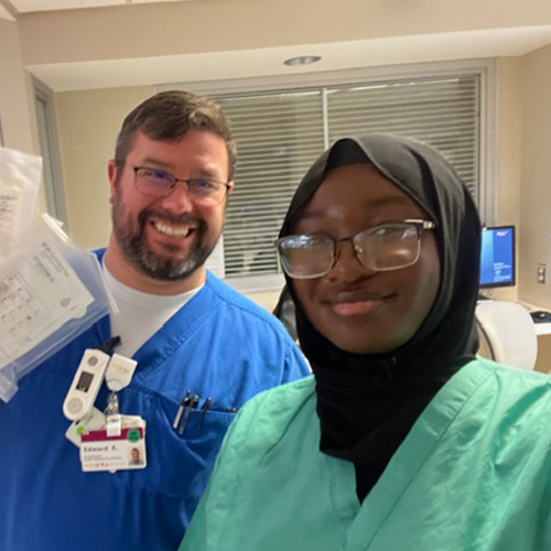 Student in green medical scrubs takes a selfie with a smiling health care worker in a hospital room.