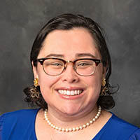A dark-haired white woman with glasses in a blue blouse with a pearl necklace