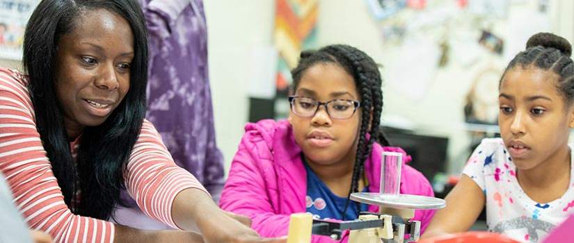 African American STEM teacher in classroom pointing to a scale with a cylinder on it. Three African American female students look on.