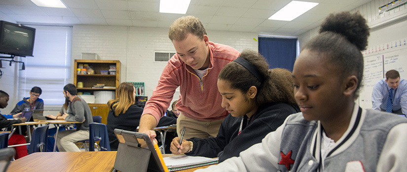 A teacher points to a tablet that a student is working on while another student smiles in the foreground.