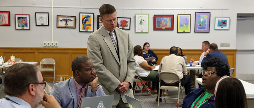 standing man talks with several seated people in busy conference room