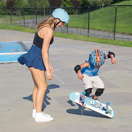 Chase Barclay teaches a young boy how to skateboard at an outdoor skate park, guiding him as he practices a trick.