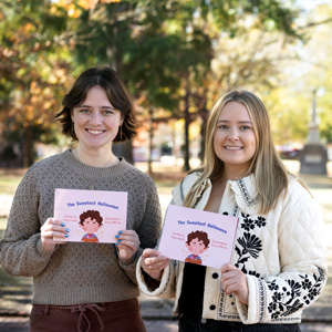 two women hold a book in an outdoor setting with trees in the background