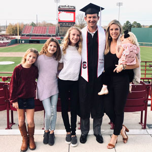 Adam Everett smiles at the baseball stadium with his family. He is wearing his graduation robe and mortarboard.
